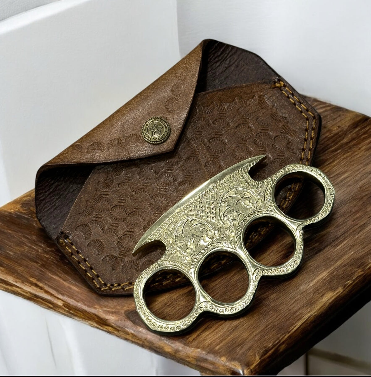 "Handmade engraved brass knuckle with intricate designs, displayed alongside its belt loop leather case."

